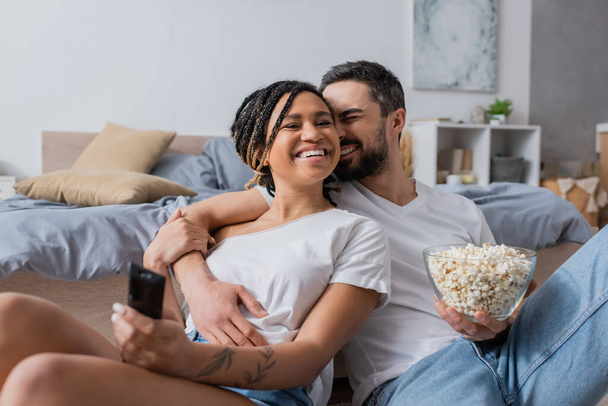 bearded man with bowl of popcorn embracing carefree african american woman with tv remote controller in bedroom at home - Fotó, kép