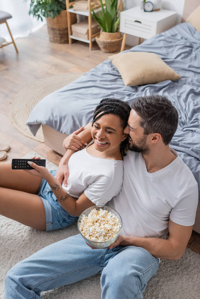 high angle view of bearded man with popcorn hugging joyful african american woman with tv remote controller on floor in bedroom - Zdjęcie, obraz