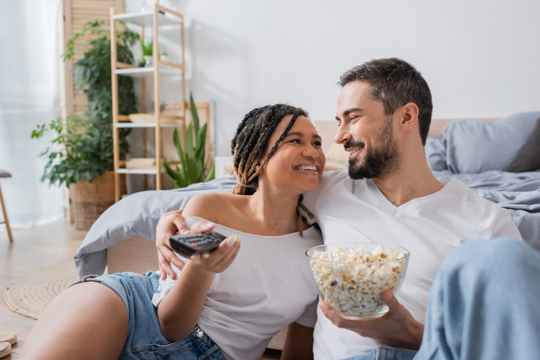 young african american woman with tv remote controller and bearded man with popcorn smiling at each other in bedroom at home - Foto, immagini