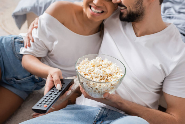 vista parziale di gioiosa coppia interrazziale con ciotola di popcorn e telecomando tv in camera da letto a casa - Foto, immagini