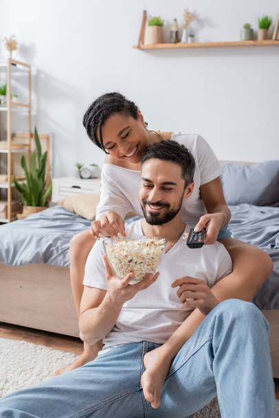 happy bearded man with bowl of popcorn sitting on floor in bedroom near african american girlfriend with tv remote controller - Foto, Bild