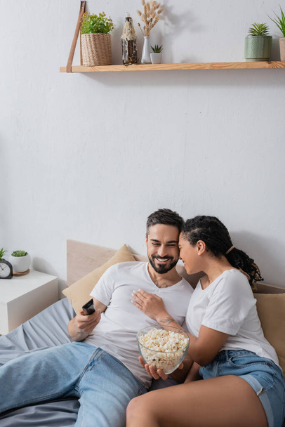 happy african american woman hugging smiling bearded man with tv remote controller and bowl of popcorn on bed at home - Fotó, kép
