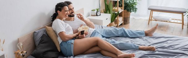 barefoot and carefree interracial couple eating popcorn and watching movie on bed at home, banner - Photo, image
