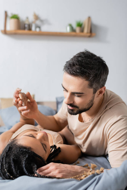 bearded man in t-shirt holding hand of passionate african american woman lying on bed at home - Foto, Bild