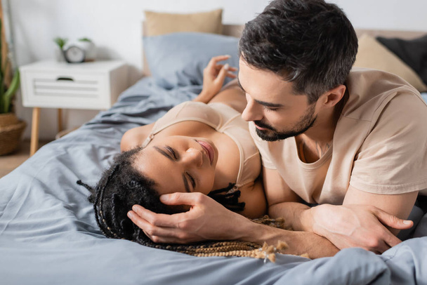 brunette bearded man touching hair of sexy african american woman in lingerie lying on bed at home - Foto, Imagem
