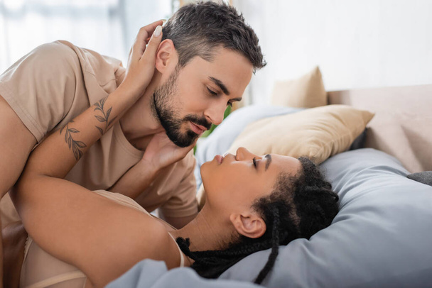 vue latérale de l'homme barbu en t-shirt près de séduisante femme afro-américaine couchée sur le lit à la maison - Photo, image