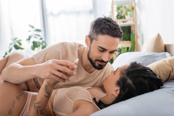 smiling bearded man holding hand of tattooed african american woman in sexy lingerie lying on bed at home - Foto, imagen