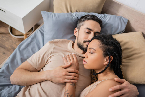 top view of bearded man with closed eyes hugging sensual african american woman while lying on bed at home - Foto, afbeelding