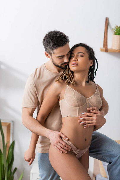 brunette man in t-shirt and jeans embracing hot african american woman in sexy lingerie in bedroom at home - Foto, Imagem