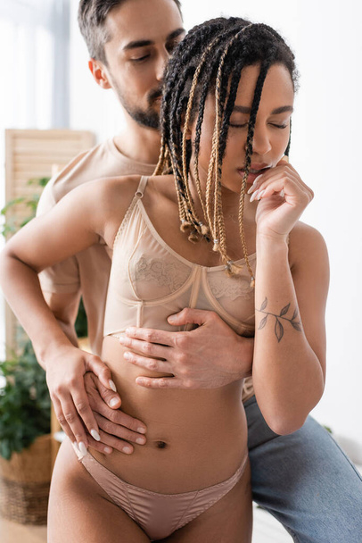 blurred man embracing sexy african american woman in underwear standing with closed eyes in bedroom at home - Foto, Imagen