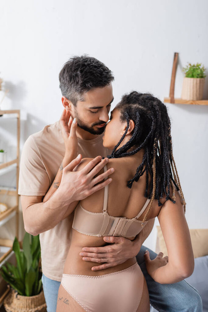 brunette man in t-shirt kissing passionate african american woman with dreadlocks in bedroom at home - Valokuva, kuva