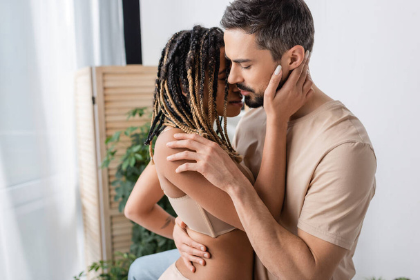 side view of bearded man in t-shirt hugging young and sexy african american woman with dreadlocks in bedroom at home - 写真・画像