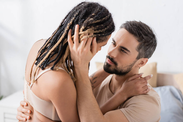 Esmer sakallı adam, yatak odasında rastalı saçlı seksi Afrikalı Amerikalı kadını kucaklıyor. - Fotoğraf, Görsel