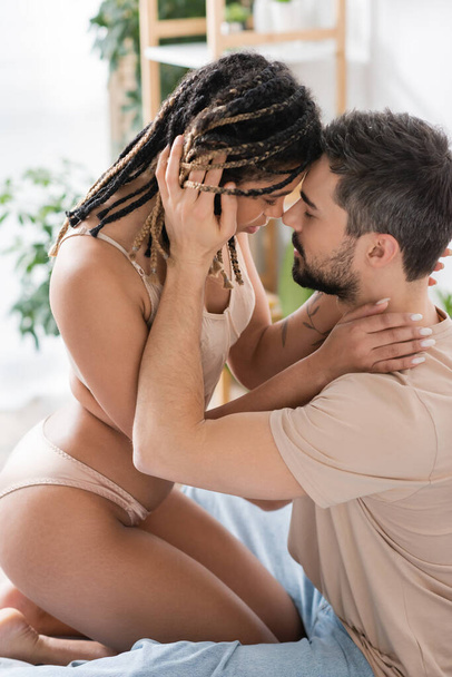側ビューの情熱的なアフリカ系アメリカ人女性でセクシーランジェリー近くブルネット男でtシャツでベッドルームに自宅 - 写真・画像