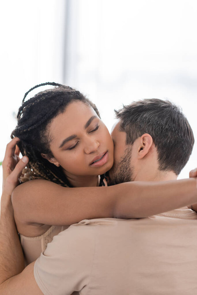 sexy african american woman with closed eyes embracing brunette man at home - Foto, imagen