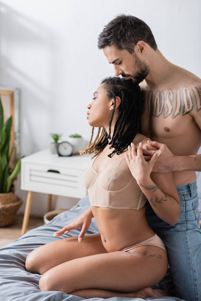 side view of shirtless tattooed man hugging young african american woman in sexy lingerie in bedroom at home - Foto, Imagem