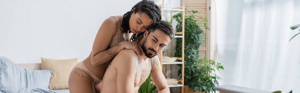 tatoué et sexy afro-américaine femme étreignant homme torse nu barbu dans la chambre à coucher à la maison, bannière - Photo, image