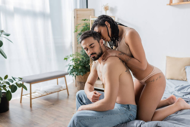 sexy africaine américaine femme en sous-vêtements séduire homme barbu torse nu en jeans dans la chambre moderne - Photo, image