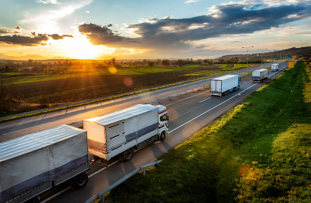 Transport samochodów ciężarowych z przyczepami na asfaltowej drodze autostradowej w krajobrazie wiejskim o zachodzie słońca - Zdjęcie, obraz