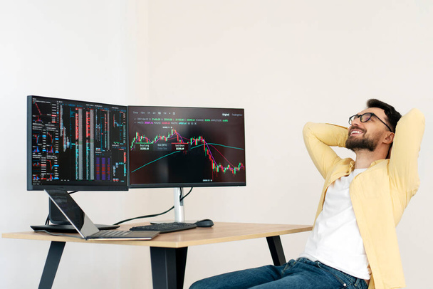Handsome smiling crypto trader leaning back, relaxing, having break sitting at workplace. Successful business, online trading concept - Φωτογραφία, εικόνα