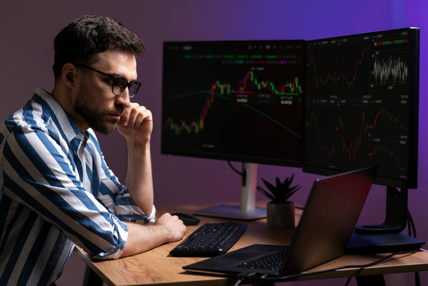Nocny portret zamyślonego handlowca patrzącego na monitor laptopa, pracującego online z wykresem crypto w nowoczesnym biurze. Handel online, udana koncepcja biznesowa - Zdjęcie, obraz