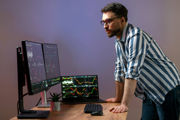 Night portrait of serious crypto trader looking at computer monitor, working online with crypto chart standing in modern office, researching and analyzing market. Technology, online trading concept - Photo, Image