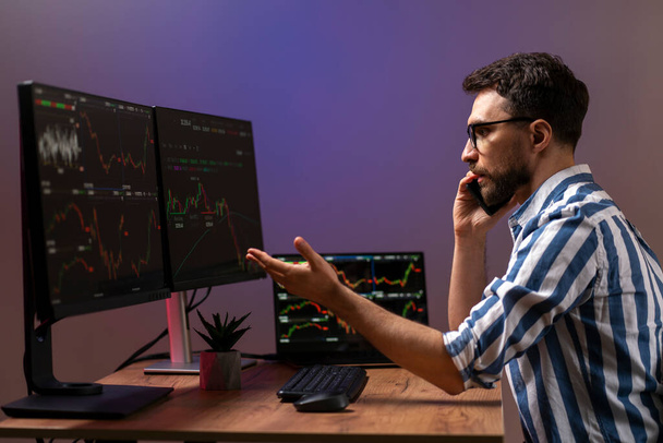 Frustrated man, crypto trader talking on mobile phone looking at computer monitor, working online with crypto chart sitting in modern office. Online trading concept - Photo, Image