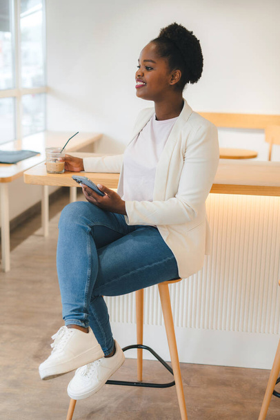 Cheerful beautiful african woman using an app in her smartphone device holding ice coffee sitting ina city cafe. Internet information. Delicious breakfast - 写真・画像