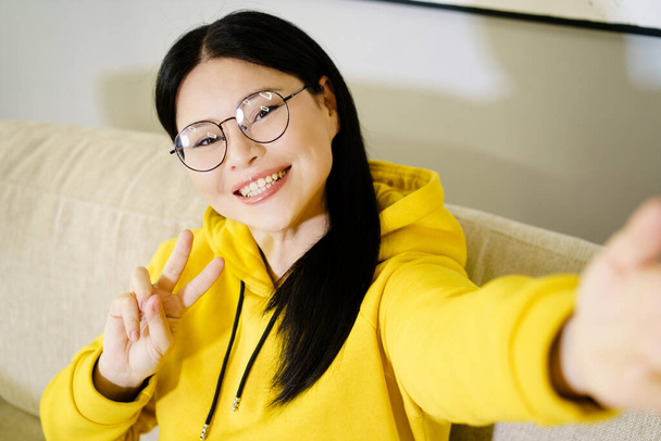 Asian woman takes self-portrait on her smartphone, smiling broadly and showing V symbol with her hand, expressing happiness and joy. High quality photo - Foto, Bild