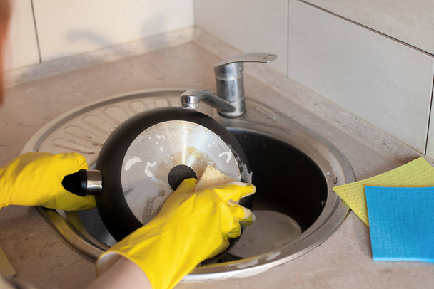 Women's hands, in yellow rubber gloves, wash a large dirty frying pan with a sponge and soap under running water under a tap. The plot is about housework and cleanliness in the kitchen. - Photo, Image