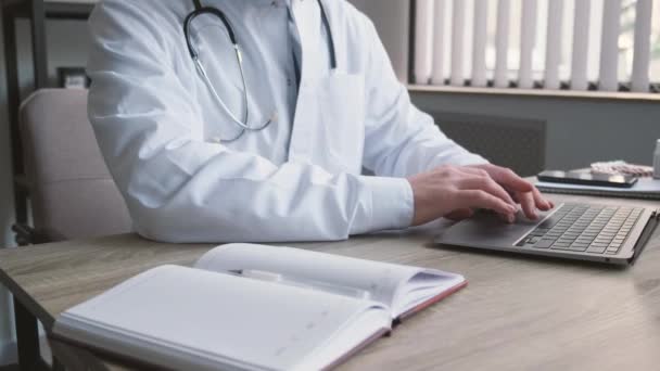 Een jonge knappe dokter werkt aan een laptop in zijn kantoor. Een moderne medische kliniek in Europa. - Video