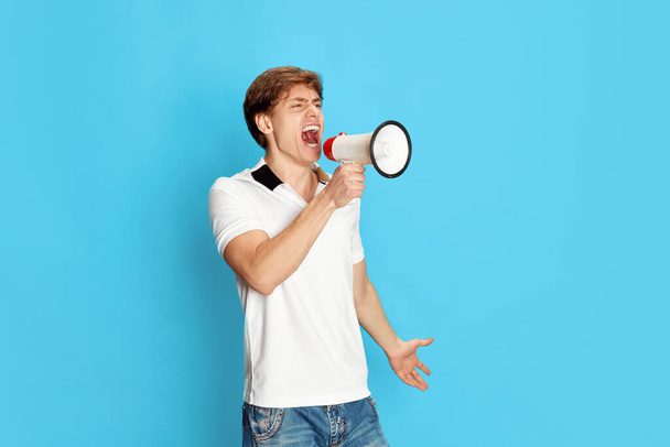 Portrait of young man, activist, emotionally shouting in megaphone against blue studio background. News, propaganda. Male casual fashion. Concept of emotions, youth, facial expression, lifestyle - Zdjęcie, obraz