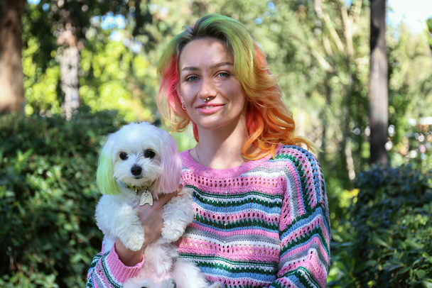 Young woman wearing colorful knitted sweater walking in a park with her maltese dog on a sunny day. Female with multicolored hair and her pup with color matched ears. Copy space, background. - Fotografie, Obrázek