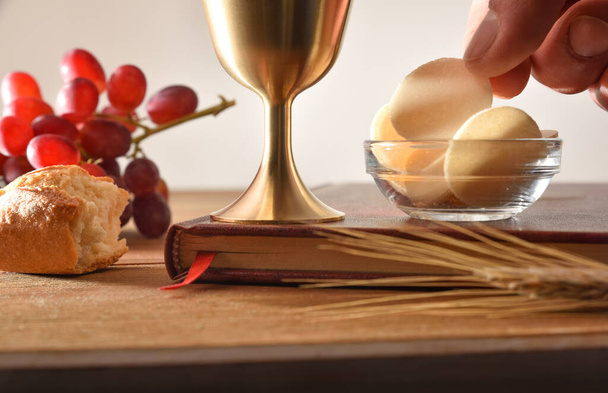 Detail of hand picking up consecrated host in container on wooden table with bible, cup, bread and grapes. Front view. - 写真・画像
