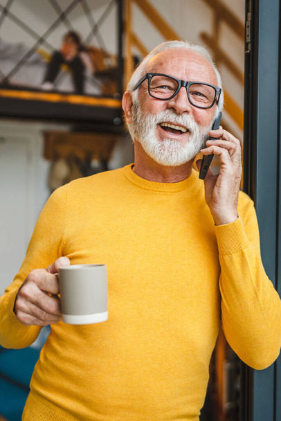 Un homme homme caucasien senior avec barbe grise debout sur le balcon à la porte dans la journée sourire heureux utiliser téléphone intelligent mobile pour faire un appel parler copie espace tenir tasse de café matin routine quotidienne - Photo, image