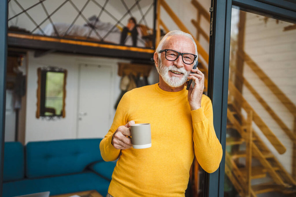 Egy férfi idősebb fehér férfi szürke szakállal áll az erkélyen az ajtóban nappal boldog mosoly használja a mobiltelefon, hogy egy hívást talk másolás hely hold csésze kávé reggeli rutin - Fotó, kép