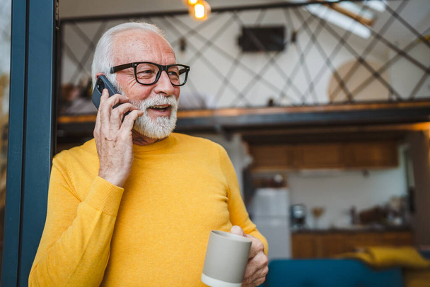 Jeden mężczyzna starszy biały mężczyzna z szarą brodą stać na balkonie w dzień szczęśliwy uśmiech korzystać z telefonu komórkowego smartfona do rozmowy telefonicznej skopiować przestrzeń trzymać filiżankę kawy rano codziennej rutyny - Zdjęcie, obraz