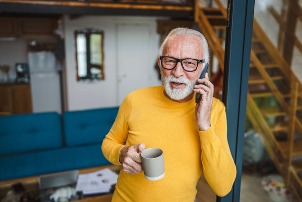 Yksi mies vanhempi valkoihoinen mies harmaa parta seistä parvekkeella ovella päivällä onnellinen hymy käyttää mobiili älypuhelin tehdä puhelun puhua kopio tilaa pidä kuppi kahvia aamulla päivittäin rutiini - Valokuva, kuva