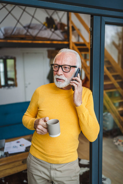 Один мужчина старший кавказский мужчина с седой бородой стоять на балконе у двери в день использовать мобильный смартфон, чтобы сделать звонок говорить скопировать пространство держать чашку кофе беспокоится вдумчивый - Фото, изображение