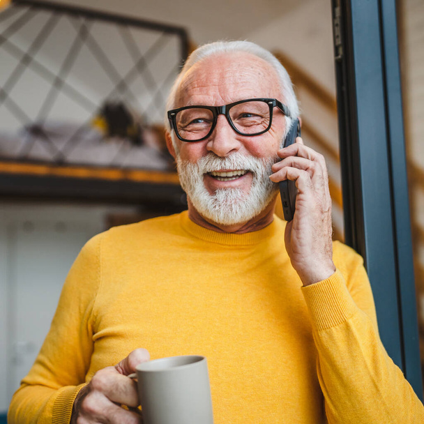 One man senior caucasian male with gray beard stand on balcony at door in day happy smile use mobile smart phone to make a call talk copy space hold cup of coffee morning daily routine - Photo, Image