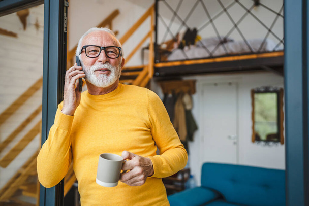 One man senior caucasian male with gray beard stand on balcony at door in day happy smile use mobile smart phone to make a call talk copy space hold cup of coffee morning daily routine - 写真・画像