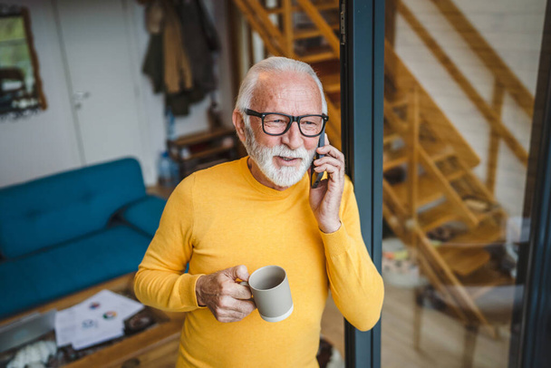 Egy férfi idősebb fehér férfi szürke szakállal áll az erkélyen az ajtóban nappal használja a mobiltelefon, hogy egy hívást talk másolás helyet tartani egy csésze kávét aggódó gondolkodó - Fotó, kép