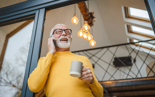 Jeden muž senior kavkazský muž s šedým vousem stojí na balkóně u dveří ve dne šťastný úsměv pomocí mobilního chytrého telefonu, aby hovor talk copy prostor držet šálek kávy ranní rutina - Fotografie, Obrázek