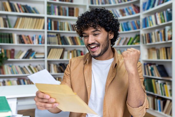 Young student received letter with good exam results and university admission confirmation, hispanic man with curly hair celebrating successful achievement holding hand up close up. - Фото, зображення