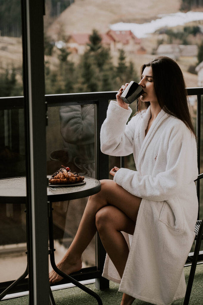 Una mujer en un albornoz blanco come un croissant y bebe café en un hotel en las montañas.Concepto de vacaciones Resort - Foto, Imagen