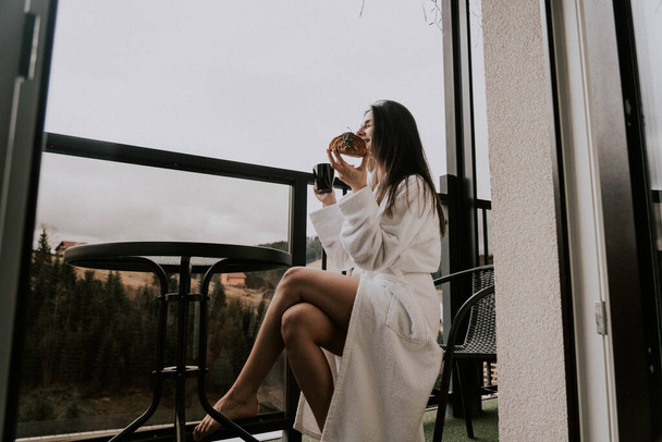 Kobieta w białym szlafroku je rogalika i pije kawę w hotelu w górach.Koncepcja wypoczynku w kurorcie - Zdjęcie, obraz