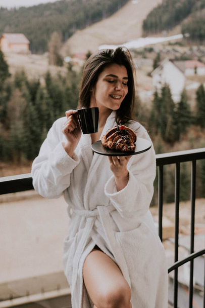Una mujer en un albornoz blanco come un croissant y bebe café en un hotel en las montañas.Concepto de vacaciones Resort - Foto, imagen