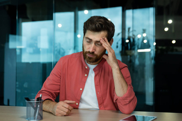 Shocked and upset young man sitting at desk in office, thoughtfully holding head with hand, closed eyes, crying, feeling pain Fired from job, salary reduction, unemployed. - Valokuva, kuva