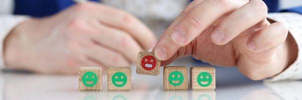 Close-up of male hand holding wooden cubes with angry emoji. Man giving negative feedback. Service rating, ranking, customer review and feedback concept - Photo, image