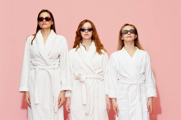Portrait of three beautiful young girls in white bathrobes and sunglasses posing with serious expression against pink studio background. Youth, face care, beauty, friendship, party, relaxation concept - Φωτογραφία, εικόνα